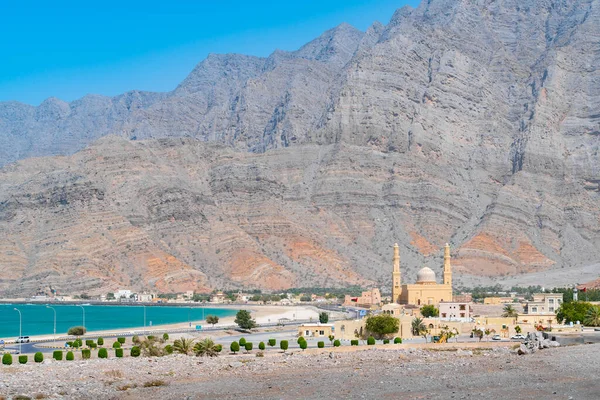 Mezquita con dos minaretes y un pequeño fuerte con el océano y altos acantilados de montaña en el fondo en un día soleado caliente. Viajar a la península de Musandam, Omán. — Foto de Stock