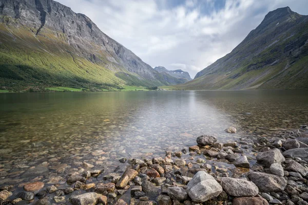 Low angle long exposure of Norwegian lake with dramatic mountains around and a small village on the other side. Cloudy day in Scandinavian landscape. Autumn in Norway. — Zdjęcie stockowe