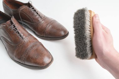 Studio shot of old, weary and little scratched cap toe lace op oxford shoes in burnished brown color, on white background. Right male hand holding a shoe brush. Old shoes before cleaning and polishing