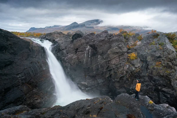 Homem em pé sobre uma rocha de cachoeira poderosa sob o céu dramático. Cachoeira Stuor Muorkke no parque nacional Stora Sjofallet, Suécia. Aventura no deserto ártico — Fotografia de Stock