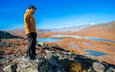 Sonbahar günü İsveç 'teki Stora Sjofallet Ulusal Parkı' nın engin arktik manzarasına bakan sarı kabuklu ceketli ve siyah pantolonlu bir erkek yürüyüşçü. Laponya dağları ve vadileri
