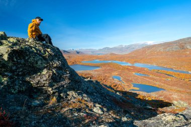 Sonbahar günü İsveç 'teki Stora Sjofallet Ulusal Parkı' nın engin arktik manzarasına bakan sarı kabuklu ceketli ve siyah pantolonlu bir erkek yürüyüşçü. Laponya dağları ve vadileri