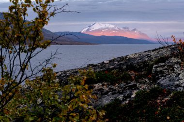 Gündoğumunun destansı ışığı kayalıkları aydınlatıyor. Buzullar arka planda kara bulutlu gökyüzü ile kaplı. Ahkka altın saat ışıklarıyla zirveye ulaştı, Stora Sjofallet Ulusal Parkı, İsveç. Kuzey Kutbu manzarası