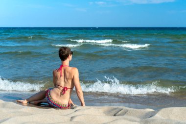 Attractive sporty short haired woman in her thirties, wearing bikini, sitting on a beach close to the sea, looking at the horizon. Concept for holiday, vacation, relaxation, health, joy, recreation