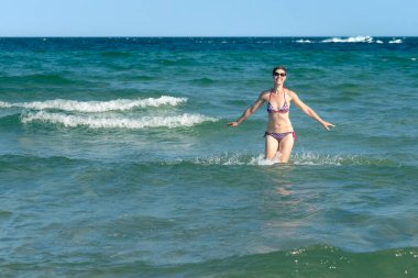 Attractive sporty short haired woman in her thirties, wearing bikini, standing in the blue sea with her arms spread on a sunny summer day. Concept for holiday, vacation, relaxation, mental health,joy