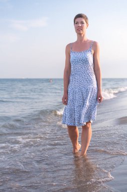 30 'larında kısa saçlı çekici bir kadın açık yaz elbisesi giyiyor. Sahilde yalınayak yürüyor. Arka planda deniz var. Tatil, rahatlama, rahatlama, fiziksel ve zihinsel sağlık kavramı