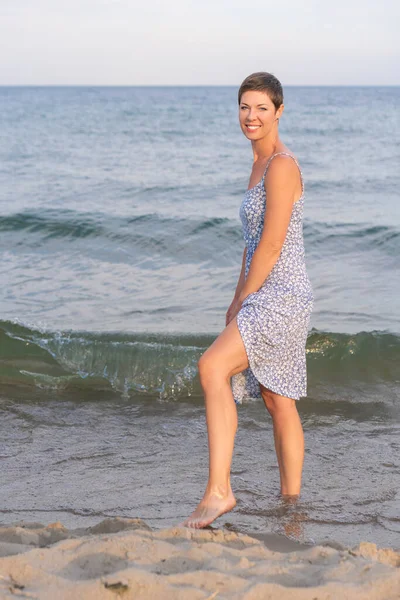 Attractive short haired woman in her thirties wear in light summer dress walks barefoot on the beach with sea in the background. Concept for vacation, leisure, relaxation, physical and mental health