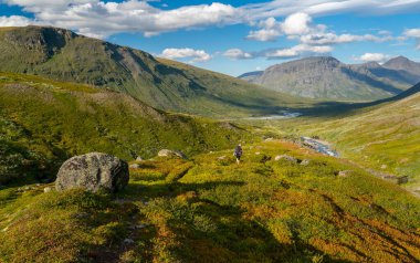 Uzak Kuzey Kutbu 'ndaki dağ vadisinde güneşli yaz gününde ağır sırt çantası ve teçhizatlı kadın yürüyüşçü. Arkasında Laddebakte dağı, Sarek Ulusal Parkı, Lapland, İsveç olan Noajdevagge vadisi..