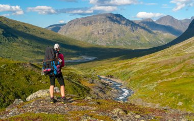 Uzak Kuzey Kutbu 'ndaki dağ vadisinde güneşli bir yaz gününde ağır sırt çantalı ve teçhizatlı kadın yürüyüşçü. Arkada Laddebakte dağı, Sarek Ulusal Parkı, Lapland, İsveç