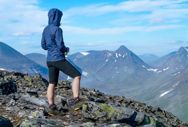 Dağın tepesinden geniş arktik dağ manzarasına bakan mavi ceketli kadın yürüyüşçü. Naite Tepesi, Sarek Ulusal Parkı, İsveç. Laponya 'da yürüyüş. Kuzey Kutbu 'nda açık hava macerası