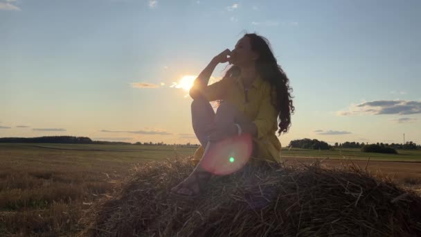 4k Silhouet van droomvrouw in een jurk zittend op een hooiberg tegen de zon. — Stockvideo
