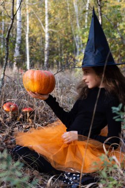 Küçük cadı balkabağı tutuyor, ona bakıyor ve gülümsüyor. Cadılar Bayramı konsepti.