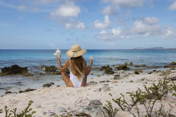 戴着草帽的女孩坐在海滩上，手里拿着大贝壳. — 图库照片