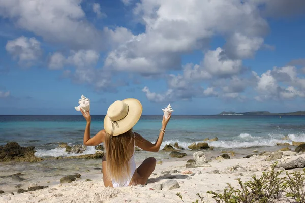 戴着草帽的女孩坐在海滩上，手里拿着大贝壳。后视镜. — 图库照片