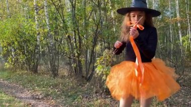 Little serious girl witch with magical stick standing on a forest path. Little girl in orange tutu skirt and black hat showing magical trick with a magic wand. Halloween concept. 4K