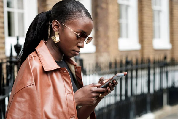 Genç siyahi kadın cep telefonunu açık havada kullanıyor. Moda, güneş gözlüğü, küpe ve deri ceket giyiyor..