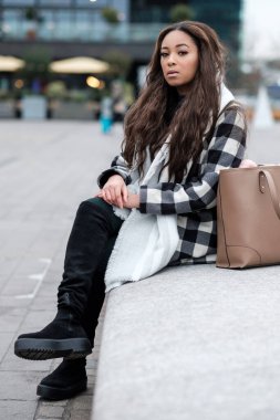 Genç siyah bir kadın taş bir bankta oturmuş kameraya bakıyor. Soğuk bir kış gününde palto, atkı ve çanta giyiyor..