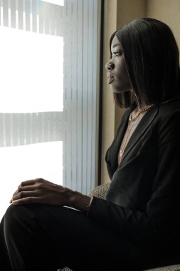 Genç siyah bir kadın model pencereden bakıyor. İş kadını kıyafeti giyiyor ve bir koltuğa oturuyor..