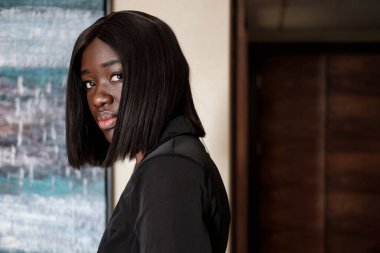 Genç siyahi kadın kameraya bakıyor. Siyah bir takım elbise giyiyor ve otel odasında..