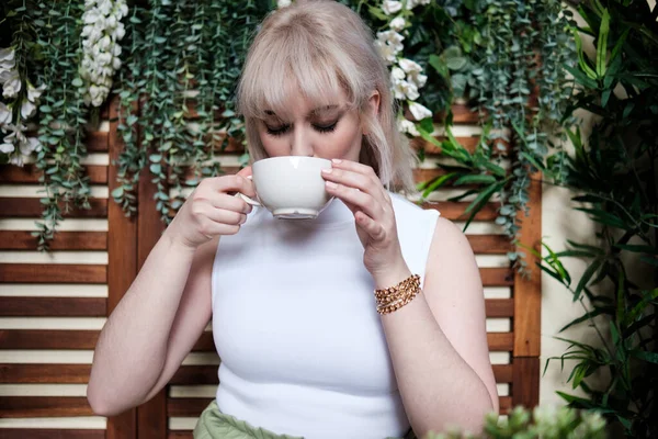 Bir kafede kahve içen çekici genç bir kadın. Beyaz bir bluz giyiyor ve arkasında bitkiler var..