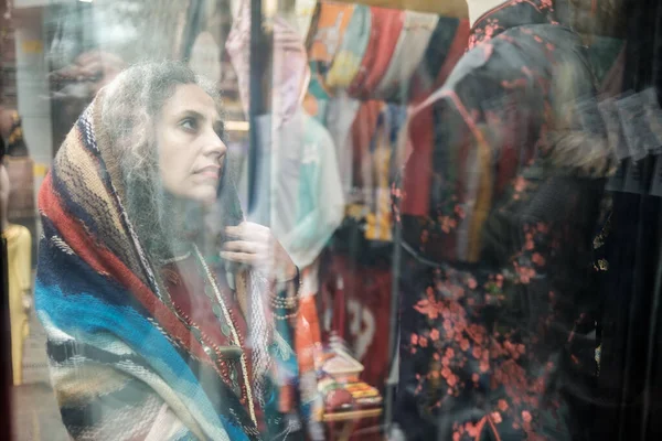 Iranian mature woman watching outfits through window shop. She is wearing a scarf on her curly and gray hair.