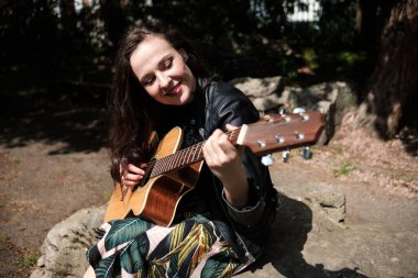 Güneşli bir bahar gününde parkta gitar çalan genç bir kadın. Gülümsüyor ve siyah deri ceket ve bahar elbisesi giyiyor..