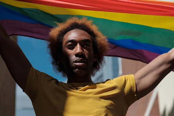Non-binary young person holding a Lgbtq flag over his head and looking away. He is between building.