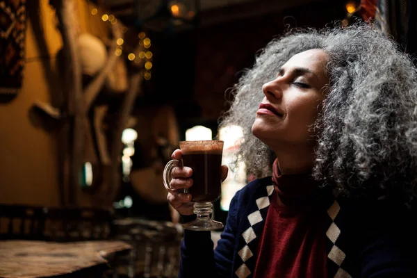 Iranian mature woman enjoying a mocha coffee in a cozy interior space. Curly grey hair.