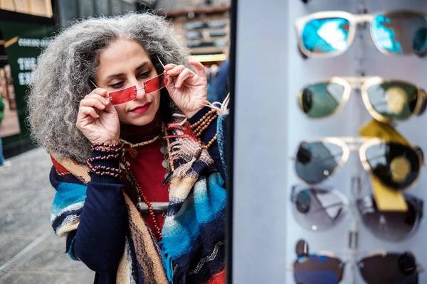 Iranian mature woman shopping sunglasses in a street market in London. She is trying sunglasses and there are many out of focus sunglasses in the foreground. Curly grey hair.