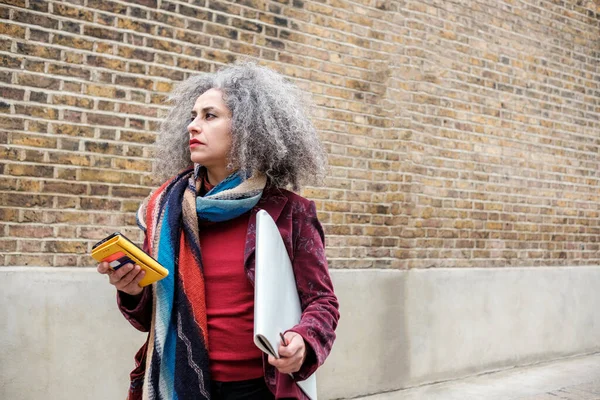 Iranian mature woman taking a sketchbook and using her phone to find a place. She is looking away and wearing colorful clothes. Grey curly hair.