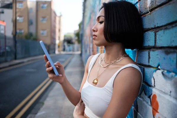 Portrait of a young female using mobile phone leaning against a wall. Side view.