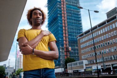 Portrait of a non-binary person in a street of London. Standing with high buildings behinds. Curly hair and yellow t-shirt.