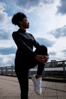 A fit black young woman is stretching close to river Thames in London. He is wearing black sport wear, curly hair.