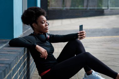 Portrait of young black woman taking a break after exercise and using phone. She is sitting on the floor, curly hair.