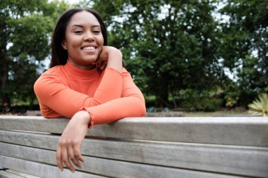 Portrait of smiling young mixed race woman sitting on a bench in a park in London and looking at camera. Orange pullover.