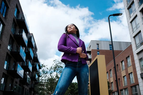 Portrait of mixed race woman model posing in a street of London. She is wearing a purple blazer and jeans.