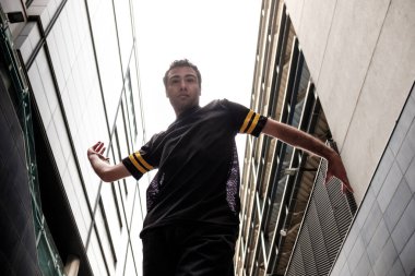A man dancer is performing an urban modern dance between some buildings in London. He is mixed race and wears black outfit.