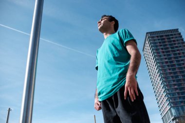 Young mixed race man is doing exercise with a bar in a London Park. High building behind.