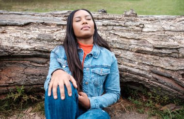A young mixed race woman is sitting on the grass and leaning against a fallen tree in a park. Her eyes are closed and she is wearing denim outfit.