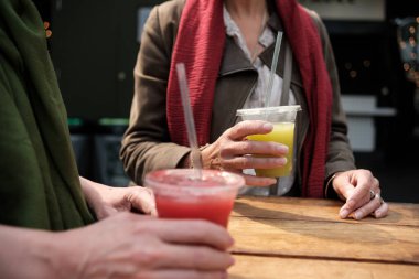 Anonymous image of two mature women hands holding juice glasses in a morning summer day. They are on a wood table and they are wearing scarfs.