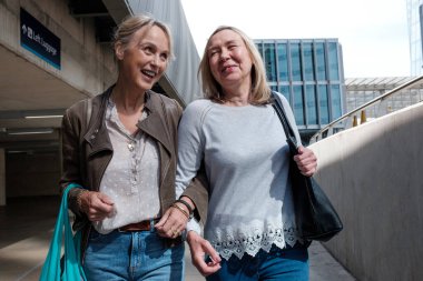 Portrait of two mature female friends walking arm laughing. They are in a modern space in London.