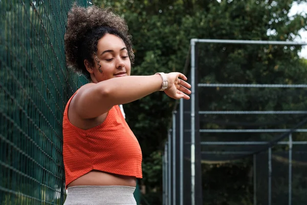 A beautiful young curvy sport model woman is checking her smart watch. Her hair is curly and she is out of cricket training court.