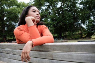 Young mixed race woman is sitting on a bench in a park and posing looking away.