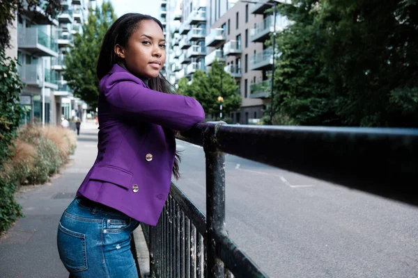 A young mixed race woman is posing in a London street looking at camera. She is leaning on a banister and she is wearing a purple blazer.