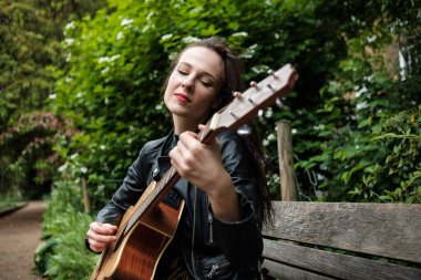 A young white woman is playing the guitar sitting on a bench's park. She is wearing a black leather jacket.