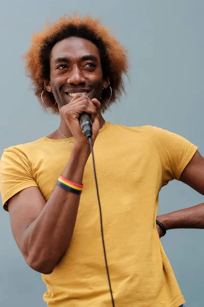 Portrait of smiling young person singing in the street holding a microphone. Wearing a lgtbq bracelet, curly hair.