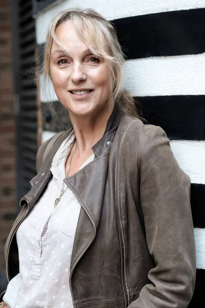 A blonde mature woman is leaning against a column and looking away. She is smiling and wearing a leather jacket.