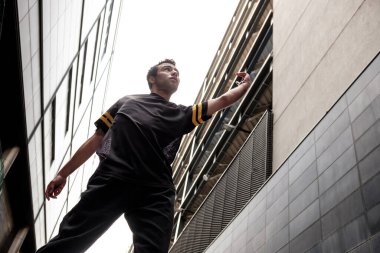 A mixed race young man is dancing between tall building in London. He is wearing black clothes.