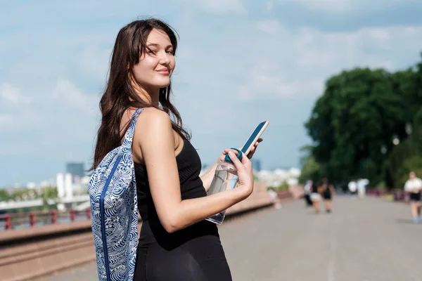 Portrait of happy young yoga woman looking at camera outdoors. She is wearing a mobile phone and a bottle of water.