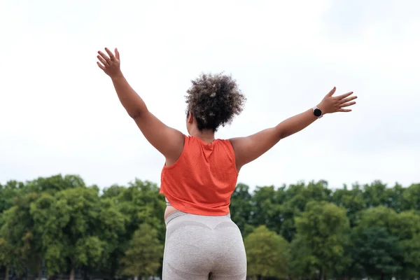 Kıvrımlı genç bir kadının zafer için kollarını açması. Konseptin üstesinden gelmek. Parkta turuncu tişört, gri tayt ve kıvırcık saç giyiyor..